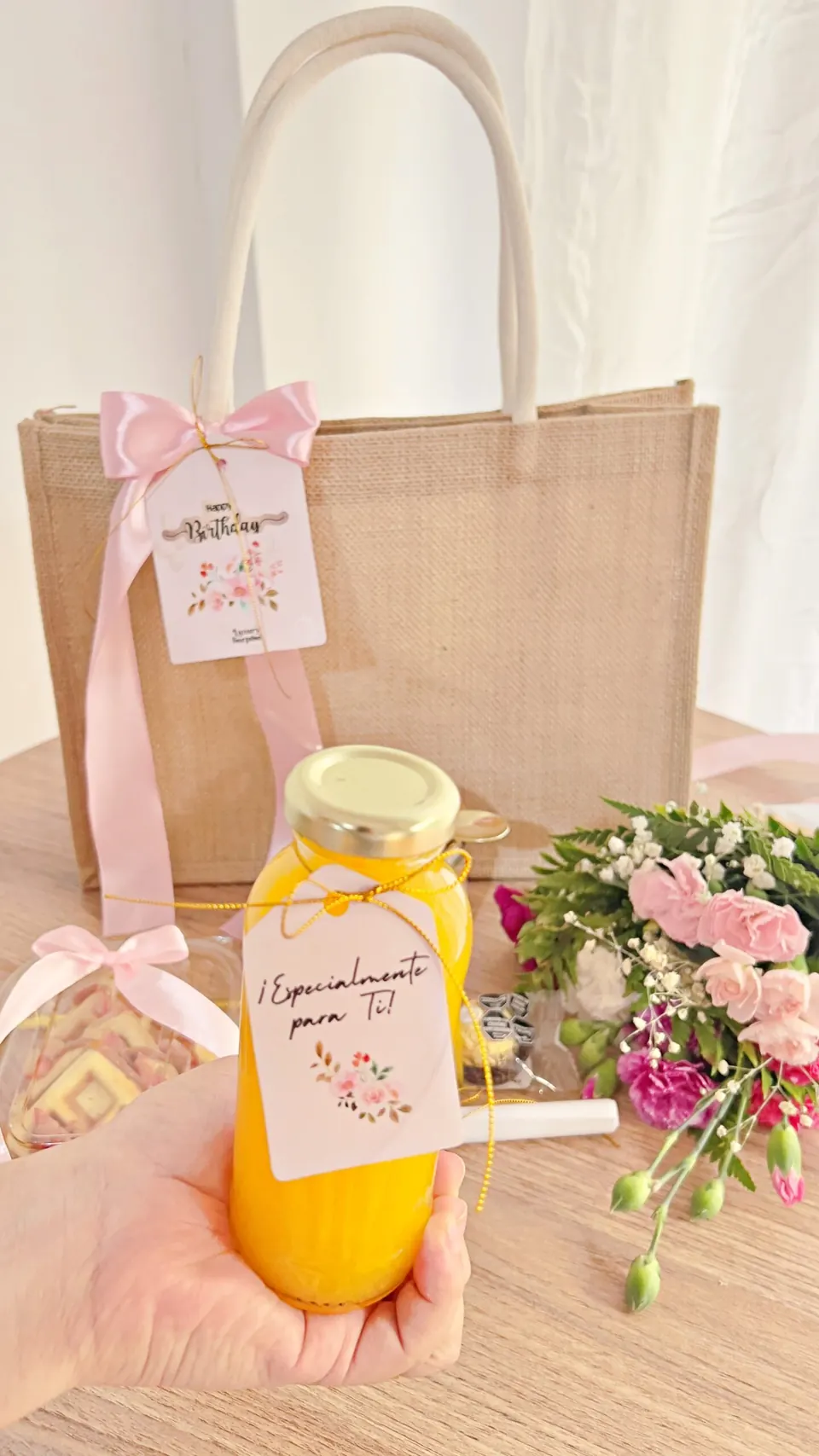 Mano sosteniendo un jugo de naranja con etiqueta personalizada junto a una cartera de regalo
